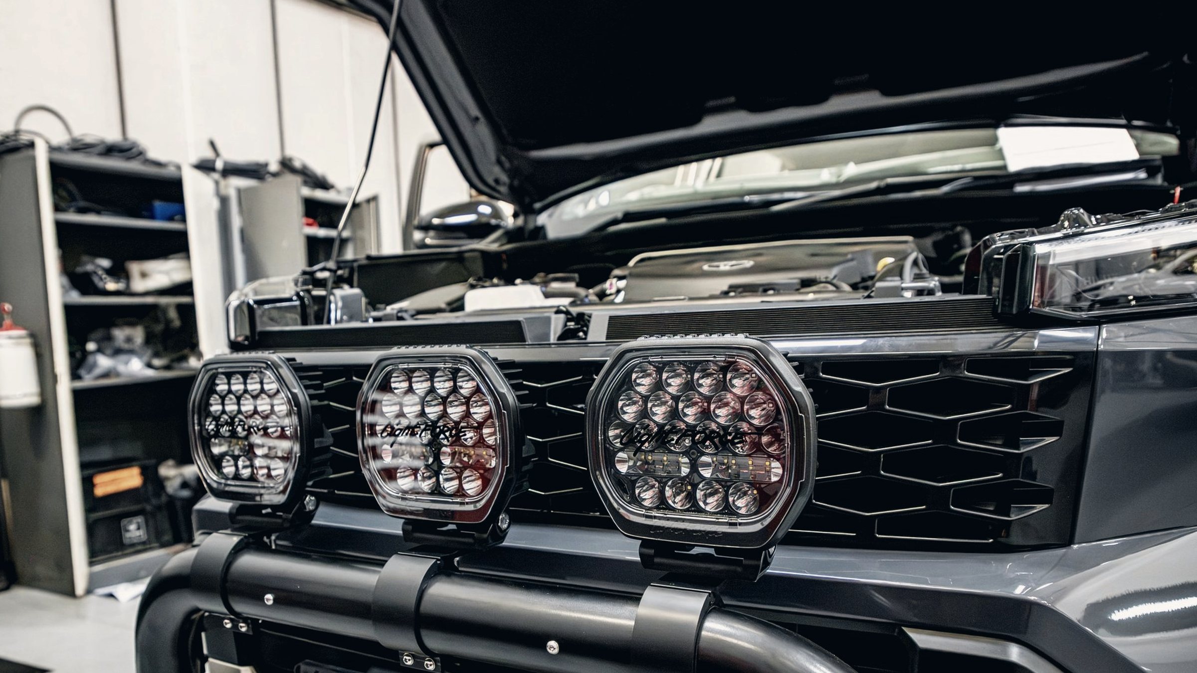 Three round LED driving lights mounted on the bull bar of a grey bakkie in a fitment bay, bonnet open.